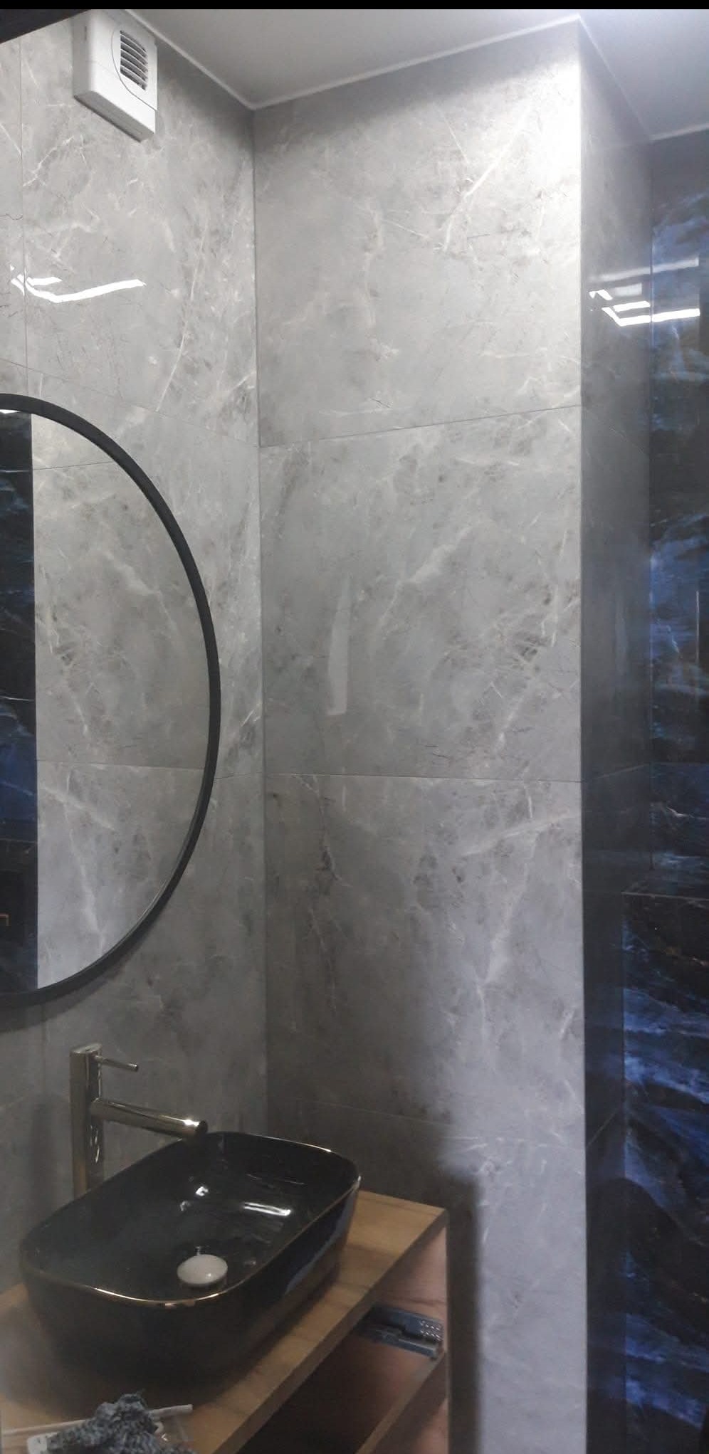 Elegancka łazienka z szarymi płytkami imitującymi marmur, okrągłe lustro z czarną ramą i czarna umywalka na drewnianym blacie. Widoczny fragment wentylacji.