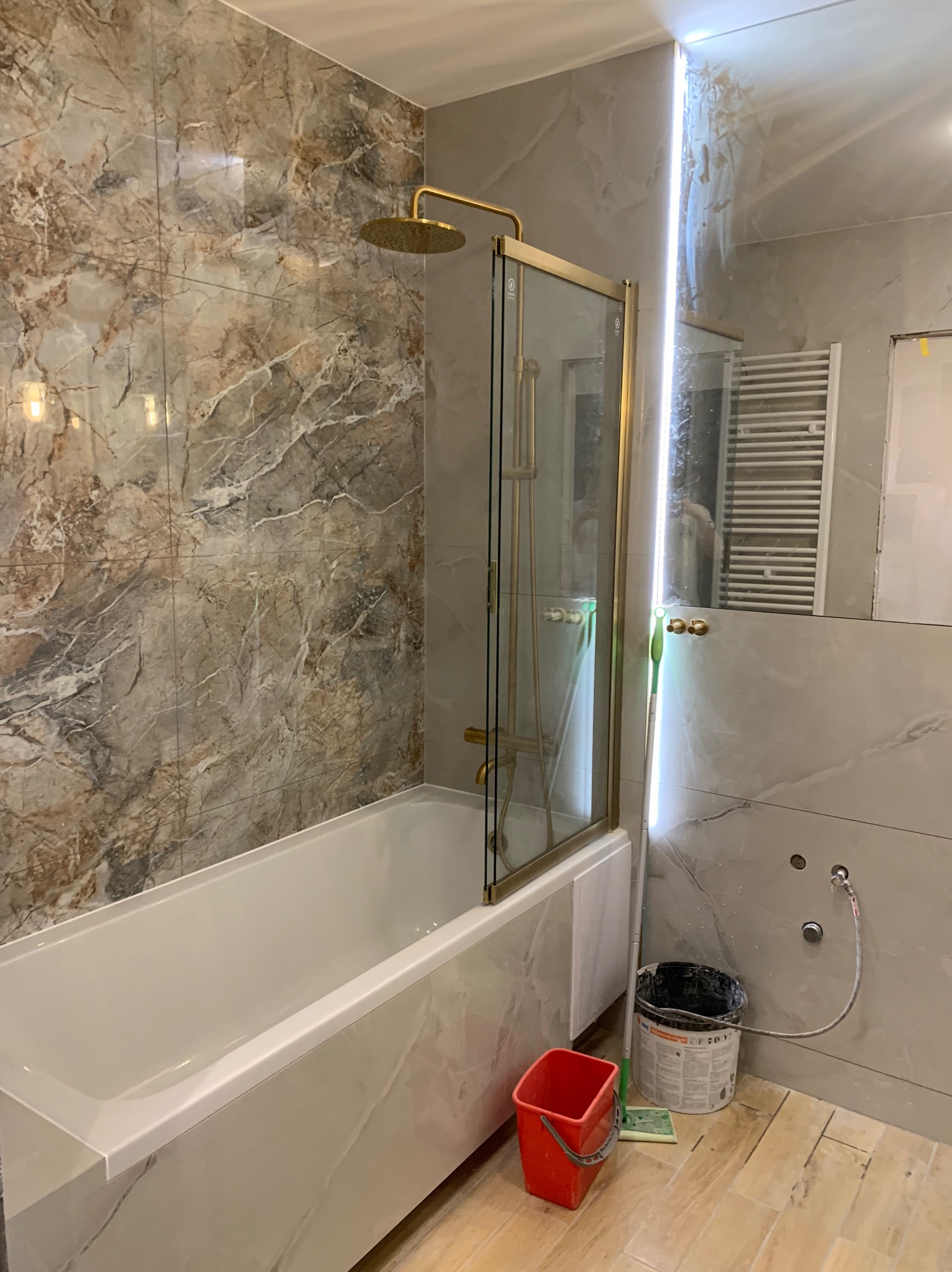 Wykończona łazienka w Żorach z wanną obudowaną płytkami imitującymi marmur, złoty deszczownica i szklany parawan. Widoczne akcesoria i wiaderka po remoncie.