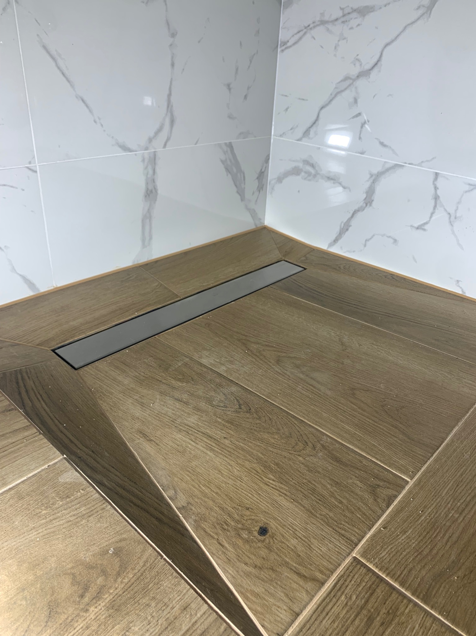 Ukończona łazienka z drewnopodobnymi płytkami podłogowymi ułożonymi w jodełkę i odpływem liniowym. Ściany wyłożone białymi płytkami imitującymi marmur.