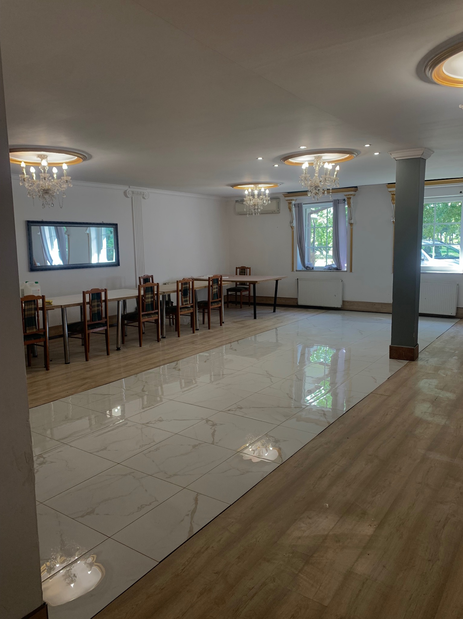 Jasne wnętrze sali z białymi płytkami podłogowymi i drewnianą podłogą, stołami i krzesłami. Dekoracyjne żyrandole i kolumna dodają elegancji.