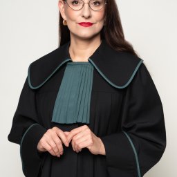 Adwokat Małgorzata Lewandowska - Portret kobiety adwokat w todze, z okularami i czerwoną szminką, na neutralnym tle. Dłonie splecione na wysokości pasa. Profesjonalny wizerunek prawnika.