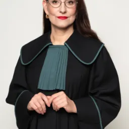 Portret kobiety adwokat w todze, z okularami i czerwoną szminką, na neutralnym tle. Dłonie splecione na wysokości pasa. Profesjonalny wizerunek prawnika.