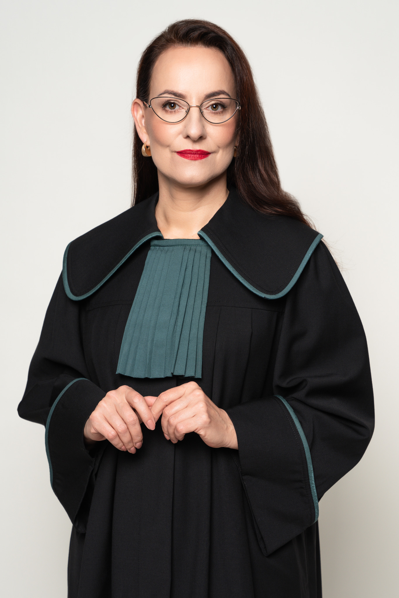 Portret kobiety adwokat w todze, z okularami i czerwoną szminką, na neutralnym tle. Dłonie splecione na wysokości pasa. Profesjonalny wizerunek prawnika.