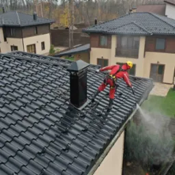 Pracownik w czerwonym kombinezonie i żółtym kasku czyści ciemny dach myjką ciśnieniową. W tle widoczne dwa domy i drzewa.