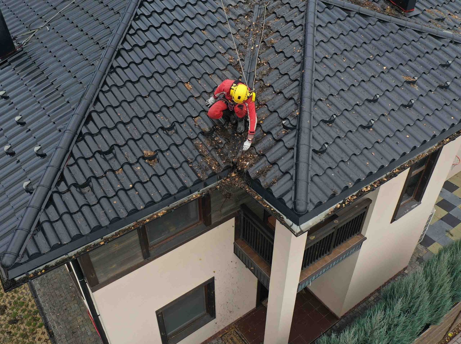 Pracownik w czerwonym kombinezonie i żółtym kasku zabezpieczony linami, z góry czyści rynnę z liści na ciemnym dachu domu.
