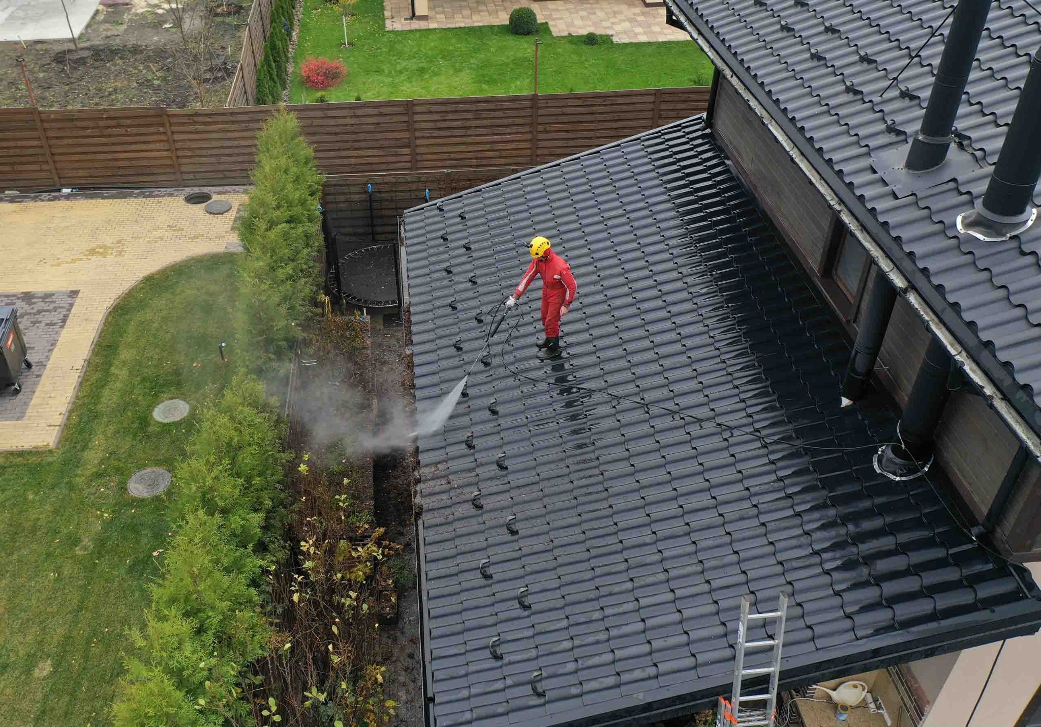Czyszczenie dachu z czarnej dachówki przez pracownika w czerwonym kombinezonie za pomocą myjki ciśnieniowej. Widok z góry na dom z ogrodem.