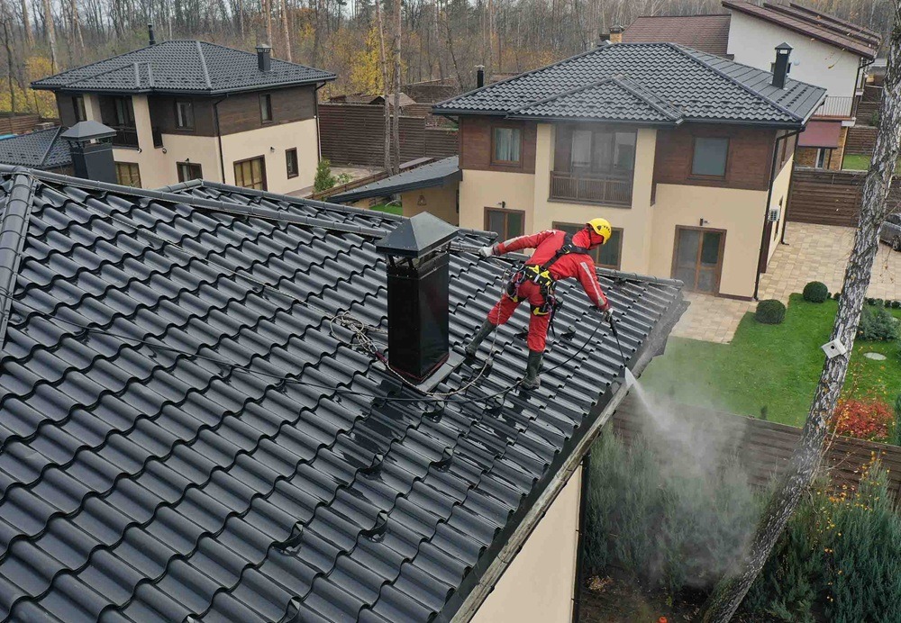 Pracownik w czerwonym kombinezonie i żółtym kasku czyści ciemny dach myjką ciśnieniową. W tle widoczne dwa domy i drzewa.