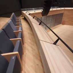 WIESŁAW OPIELA-PARKIETY.PL - Wnętrze sali widowiskowej z drewnianą podłogą i rzędem numerowanych krzeseł w odcieniach szarości. Widoczne drewniane elementy konstrukcyjne i oświetlenie.