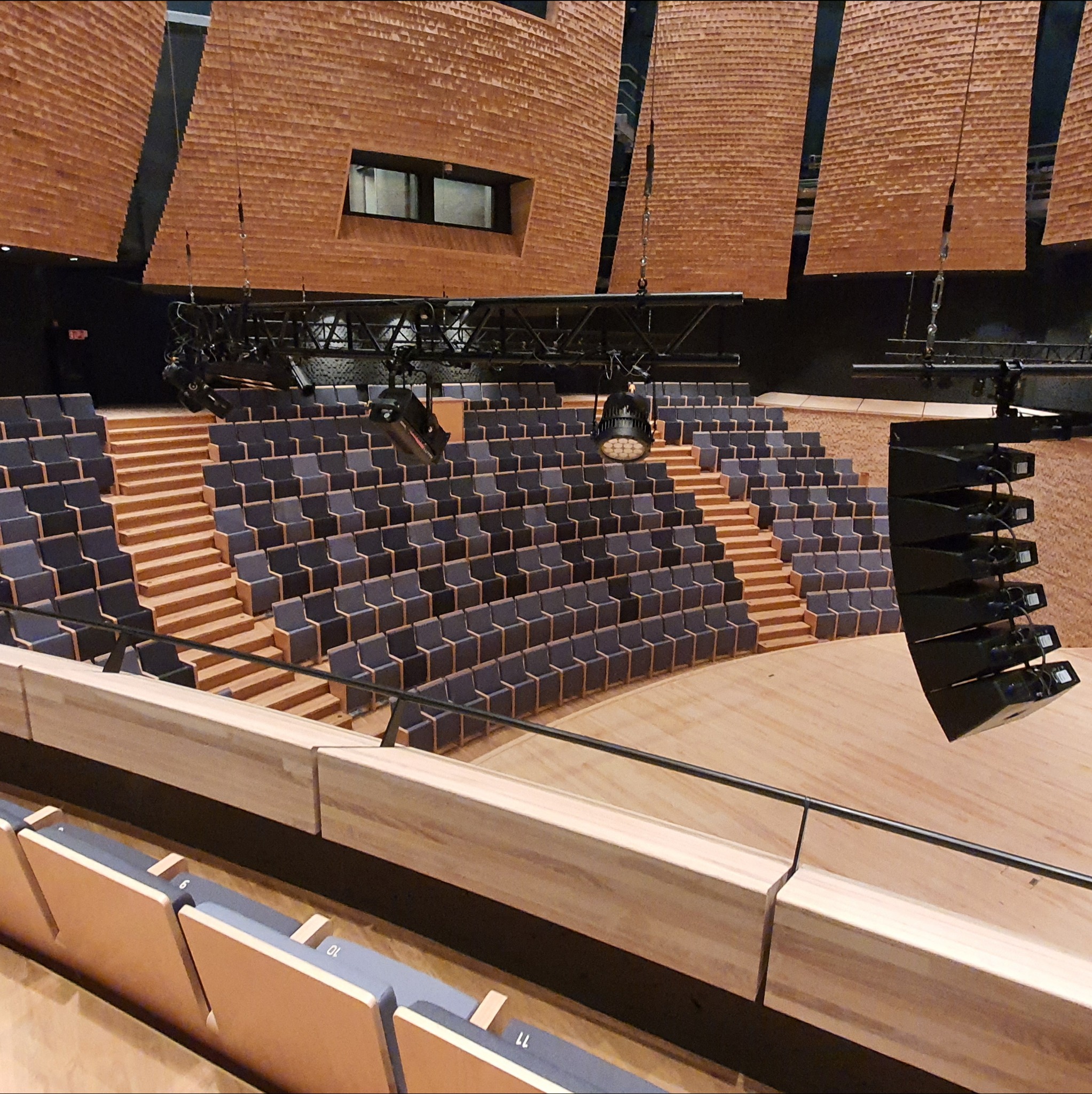 Wnętrze nowoczesnej sali widowiskowej z drewnianą podłogą, schodami i rzędami szarych foteli. Widoczne oświetlenie i elementy akustyczne na suficie.