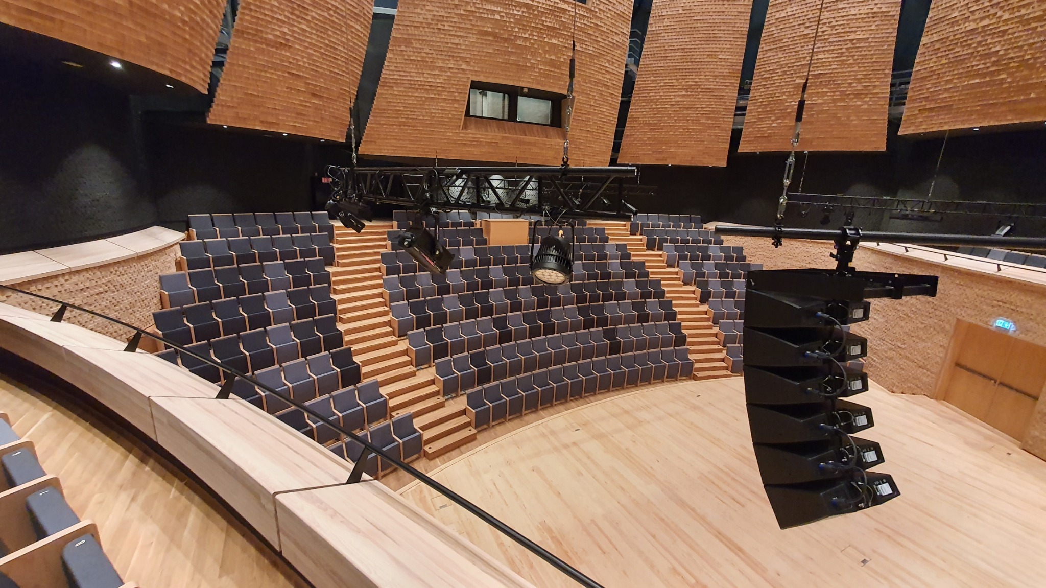 Sala koncertowa z drewnianymi schodami i rzędami krzeseł. Widoczne oświetlenie sceniczne i panele akustyczne na suficie. Całość utrzymana w ciepłej kolorystyce drewna.