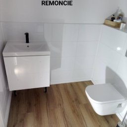Daniel Strączek - Biała łazienka po remoncie z drewnopodobną podłogą, wiszącą toaletą i szafką z umywalką. Na ścianach białe płytki. Widoczny napis 'ŁAZIENKA PO REMONCIE'.