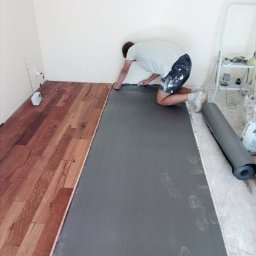 Daniel Strączek - Układanie podkładu pod panele podłogowe w jasnym wnętrzu. Widoczny fragment ułożonych paneli i rozłożony podkład. Osoba docina podkład przy ścianie.