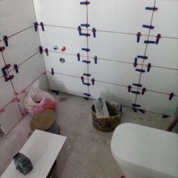 Daniel Strączek - Układanie białych płytek z niebieskimi krzyżykami dystansowymi w łazience w Prusach. Widoczne narzędzia glazurnicze i częściowo zamontowana toaleta.