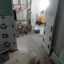 Daniel Strączek - Układanie płytek z użyciem lasera krzyżowego. W tle widoczny pracownik przygotowujący ścianę. Na podłodze narzędzia i materiały budowlane.