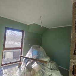 Daniel Strączek - Ściana pomalowana na zielono z białym wykończeniem, okno z drewnianą ramą, zabezpieczone meble folią i fragment ściany w trakcie prac wykończeniowych.
