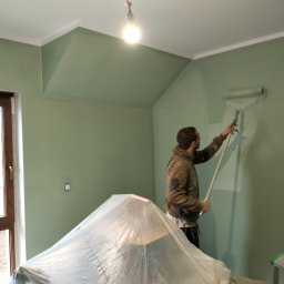 Daniel Strączek - Mężczyzna maluje ścianę na zielono wałkiem. Meble zabezpieczone folią. Widoczna lampa wisząca i okno. Ściany w odcieniu zieleni, sufit biały.