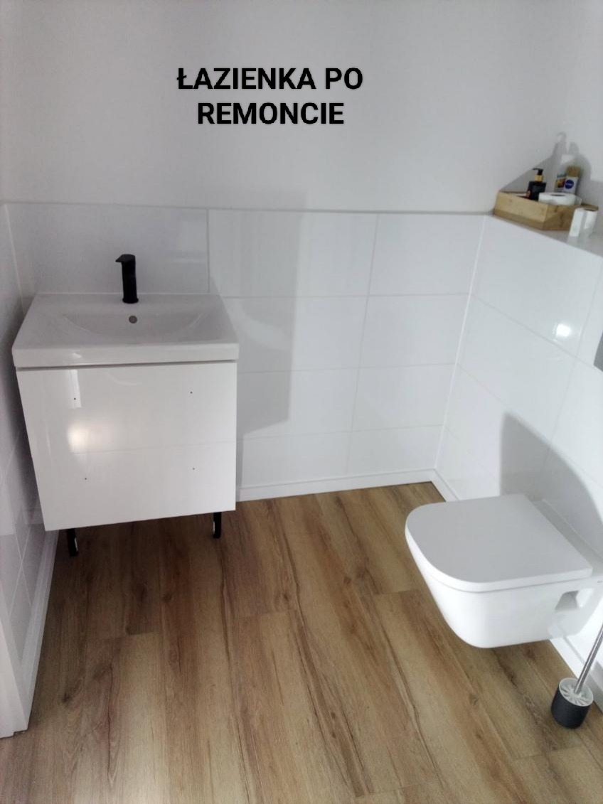 Biała łazienka po remoncie z drewnopodobną podłogą, wiszącą toaletą i szafką z umywalką. Na ścianach białe płytki. Widoczny napis 'ŁAZIENKA PO REMONCIE'.