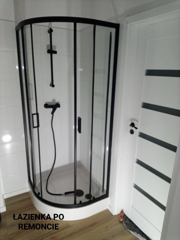 Nowoczesna kabina prysznicowa z czarnymi ramami i białą armaturą, po remoncie. Widoczna biała ściana z płytkami i drzwi z poziomymi, czarnymi paskami.