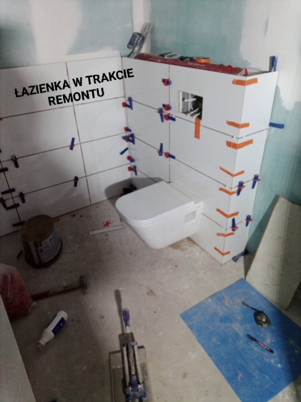 Remont łazienki w trakcie: białe płytki z krzyżykami dystansowymi, podwieszana toaleta, narzędzia glazurnicze na podłodze. Widok na etap układania płytek.
