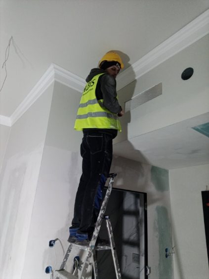Pracownik w żółtym kasku na drabinie montuje kratkę wentylacyjną w suficie z dekoracyjną listwą. Ściany w trakcie wykończenia, widoczne ślady szpachlowania.