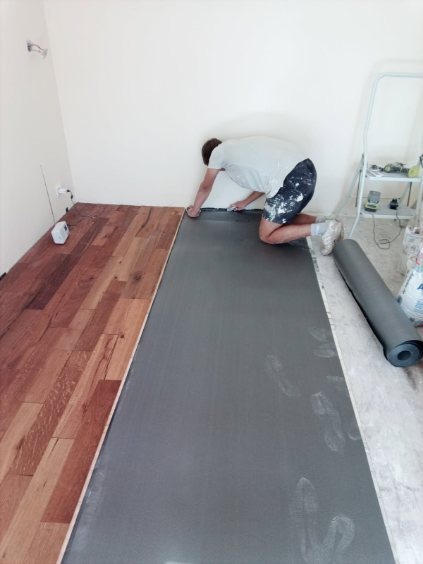 Układanie podkładu pod panele podłogowe w jasnym wnętrzu. Widoczny fragment ułożonych paneli i rozłożony podkład. Osoba docina podkład przy ścianie.