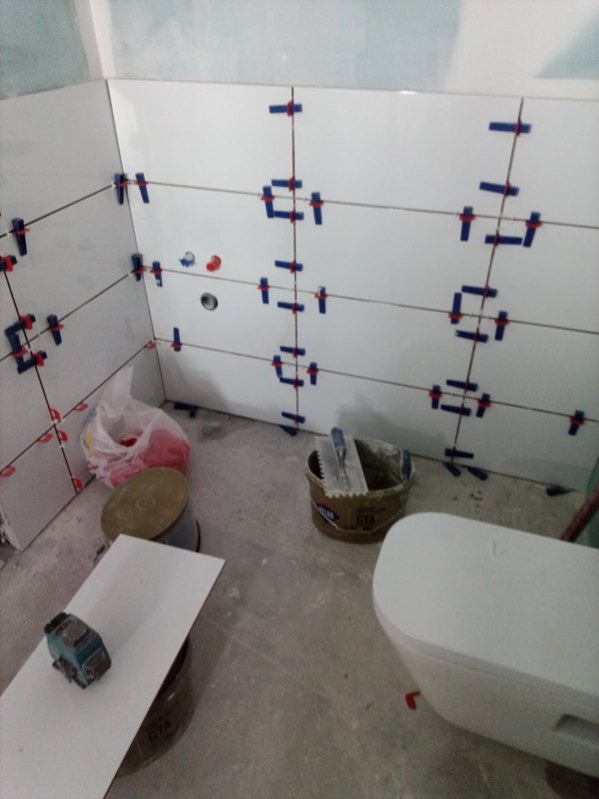 Białe płytki ścienne z niebieskimi krzyżykami dystansowymi w trakcie montażu. Widoczne narzędzia glazurnicze i fragment toalety. Ujęcie z bliska, wnętrze w trakcie prac.