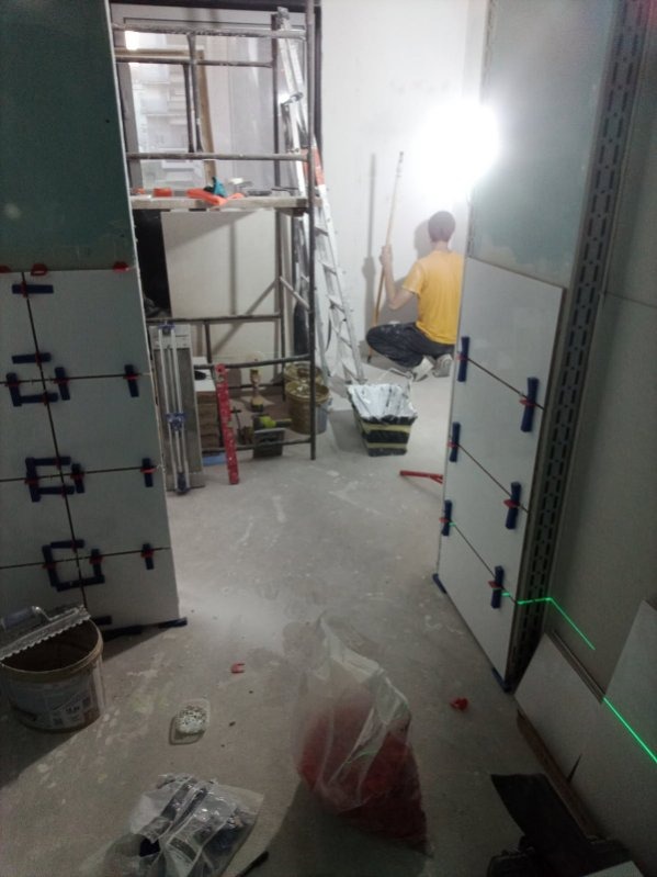 Układanie płytek z użyciem lasera krzyżowego. W tle widoczny pracownik przygotowujący ścianę. Na podłodze narzędzia i materiały budowlane.