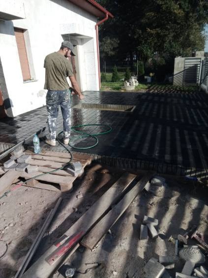 Czyszczenie kostki brukowej myjką ciśnieniową na posesji. Widoczne narzędzia i materiały budowlane. Prace brukarskie w trakcie realizacji.