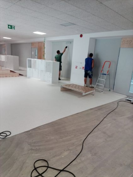 Malowanie ścian w jasnym, przestronnym wnętrzu. Dwóch malarzy przy pracy, drabina, meble i podłoga częściowo pomalowana. Widoczne kable na podłodze.