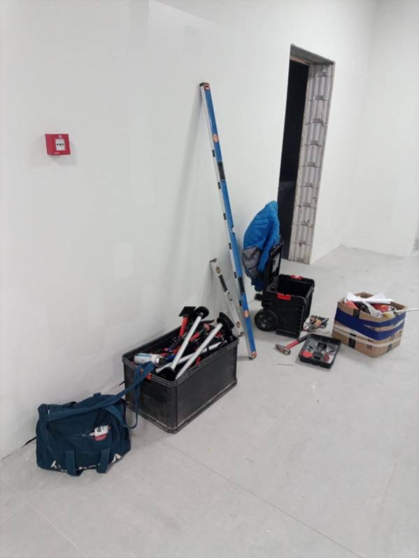 Przygotowania do prac wykończeniowych: poziomica, drabina, skrzynki z narzędziami i akcesoriami malarskimi w jasnym, odświeżonym wnętrzu z widocznym otworem drzwiowym.