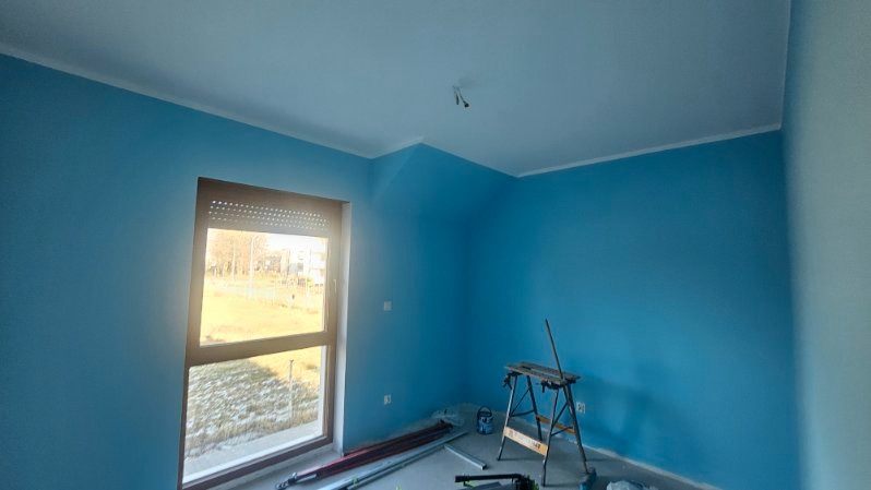 Pomieszczenie po malowaniu ścian na błękitno, widoczne narzędzia malarskie i drabina. Ściany świeżo pomalowane, okno z roletami wpuszcza światło.