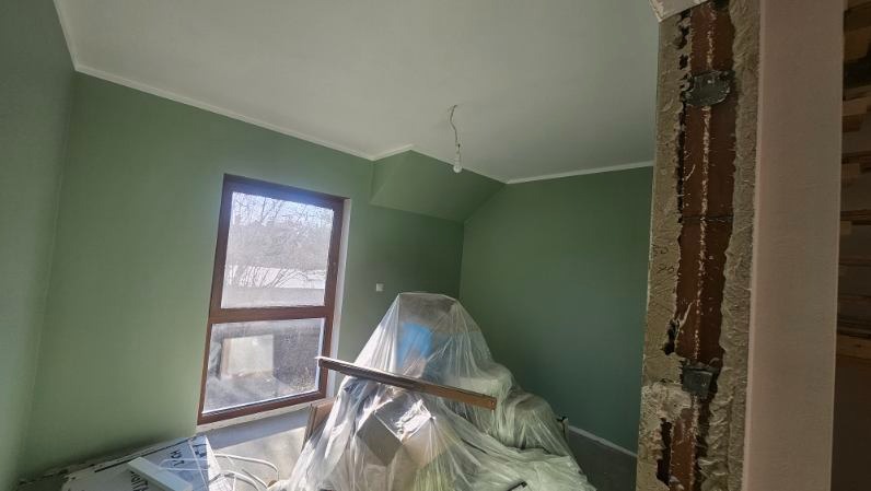 Ściana pomalowana na zielono z białym wykończeniem, okno z drewnianą ramą, zabezpieczone meble folią i fragment ściany w trakcie prac wykończeniowych.