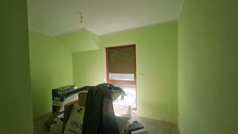 Pomalowane na jasnozielono wnętrze pokoju z oknem z brązowymi roletami. Na podłodze kartony i folia ochronna. Widoczne skośne ściany poddasza.