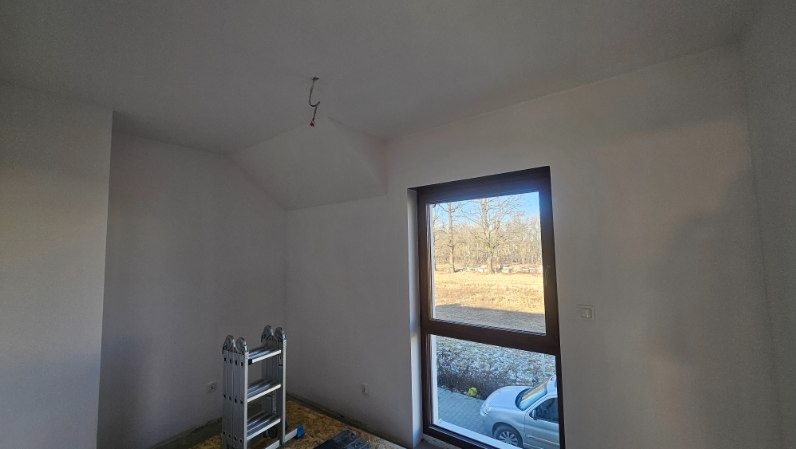 Białe ściany poddasza po malowaniu, widoczne okno z brązową ramą, drabina aluminiowa i wystający kabel ze sufitu. Widok na zewnątrz z zaparkowanym samochodem.