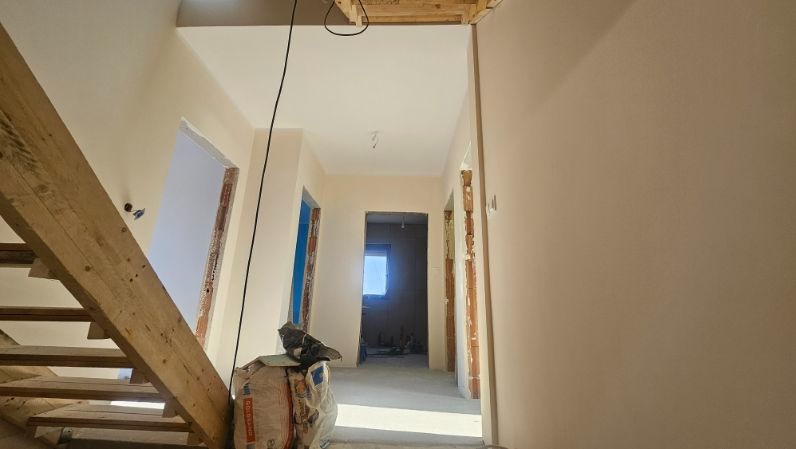 Wnętrze w trakcie prac wykończeniowych: gładkie, beżowe ściany, surowa drewniana konstrukcja schodów i worki z zaprawą. Widoczne przygotowane otwory drzwiowe.