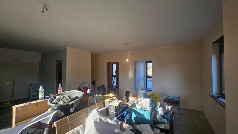 Wnętrze w trakcie remontu: widoczne narzędzia, materiały budowlane i przygotowane ściany do malowania. Częściowo wykończone pomieszczenie z oknem i drzwiami.