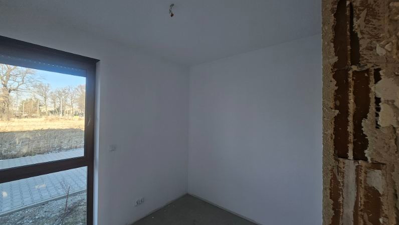 Wnętrze pokoju w trakcie prac wykończeniowych: świeżo pomalowane ściany w odcieniach bieli, surowy fragment ściany z widoczną strukturą oraz okno z widokiem na zewnątrz.