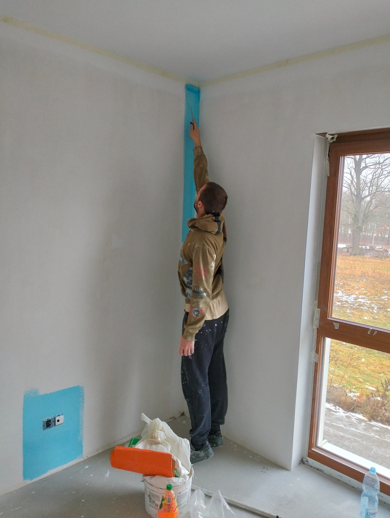 Mężczyzna maluje narożnik ściany na niebiesko, zabezpieczony taśmą malarską. Widoczne narzędzia malarskie i okno z widokiem na zewnątrz. Prace wykończeniowe w trakcie.