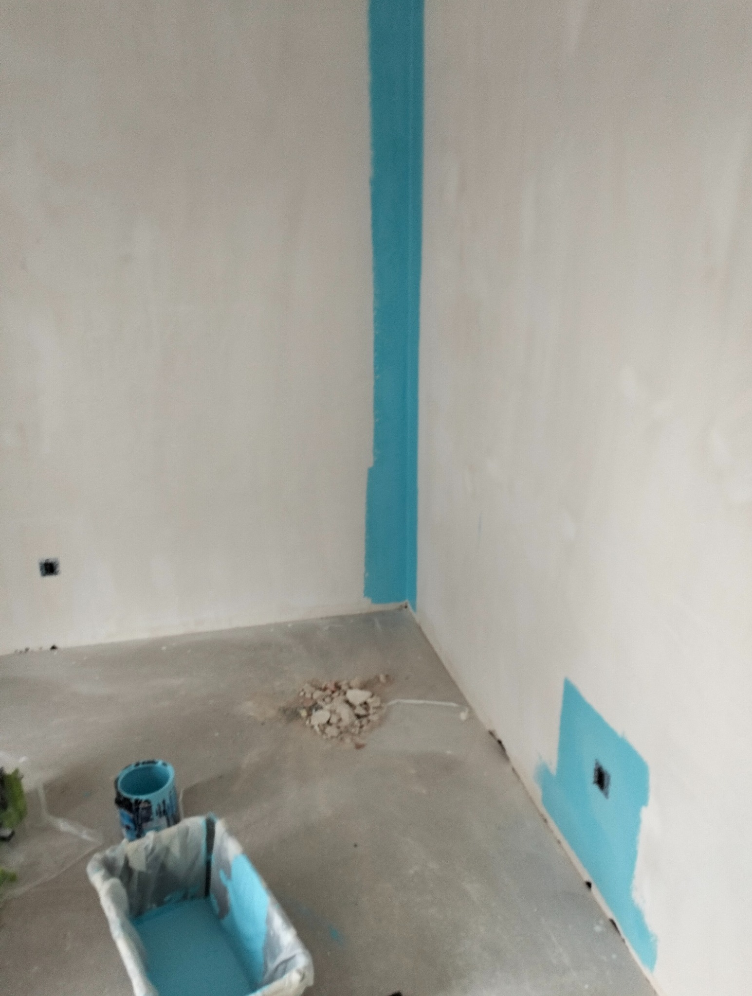 Ściana w trakcie malowania na biało z akcentami błękitu wokół narożnika i gniazdka, widoczne narzędzia malarskie i resztki gruzu na podłodze.