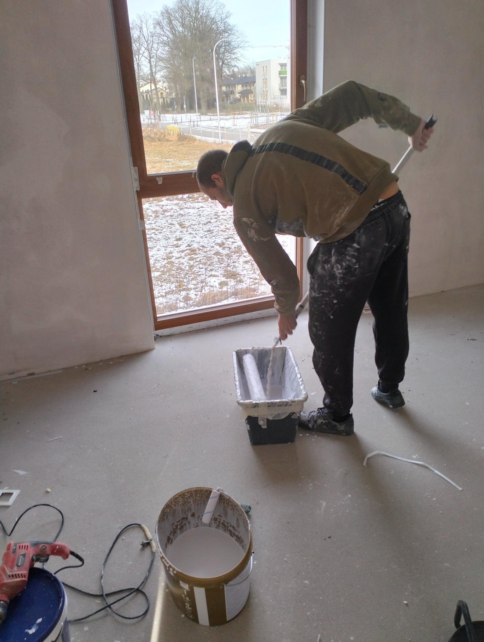 Mężczyzna w trakcie malowania ściany wałkiem w nowym budynku. Widoczne narzędzia malarskie i wiadro z farbą. Za oknem zimowy krajobraz.