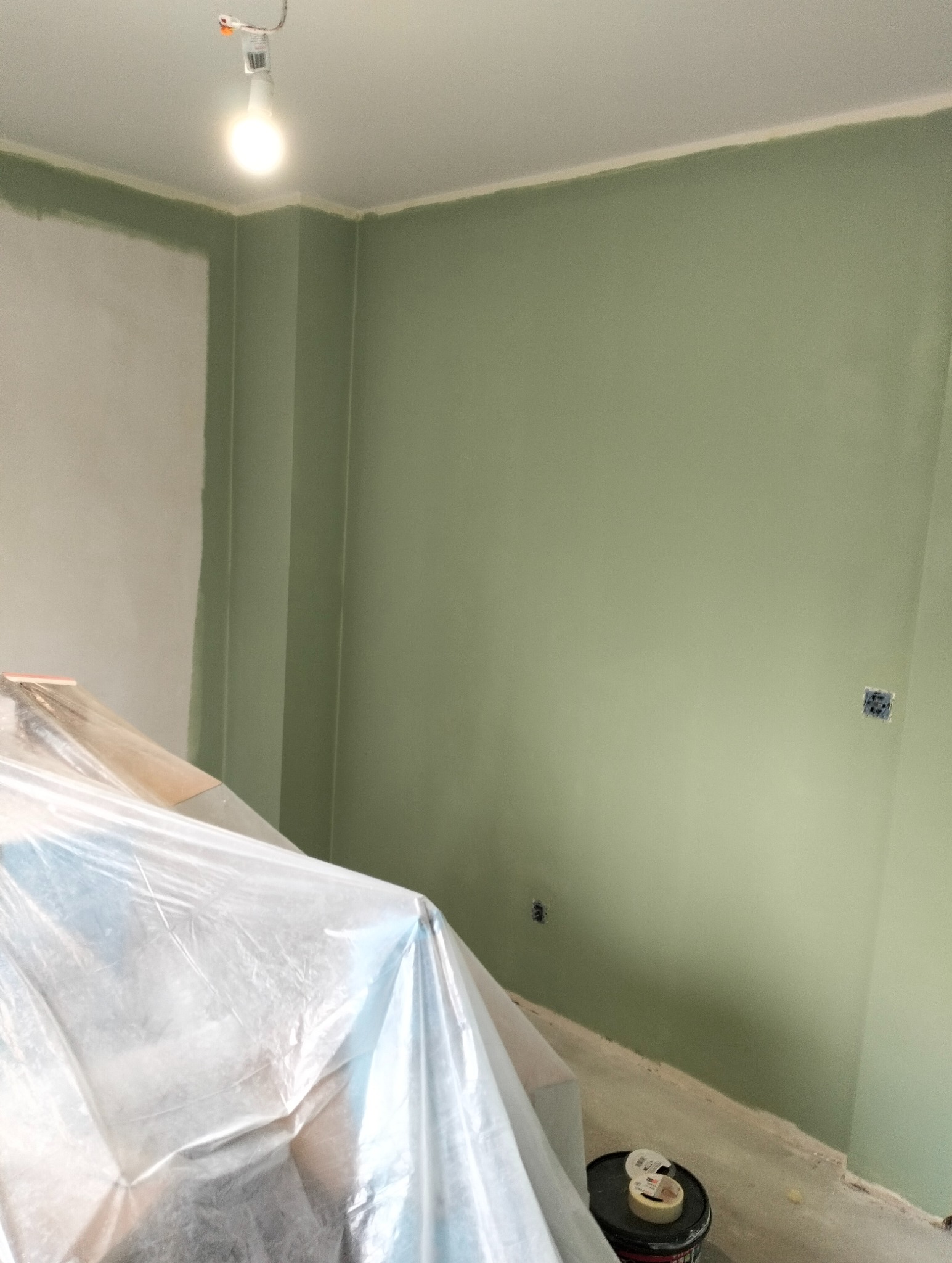 Świeżo pomalowane na zielono ściany w pokoju, zabezpieczone meble folią malarską, widoczny kubeł z farbą i taśma malarska. Oświetlenie żarówką.