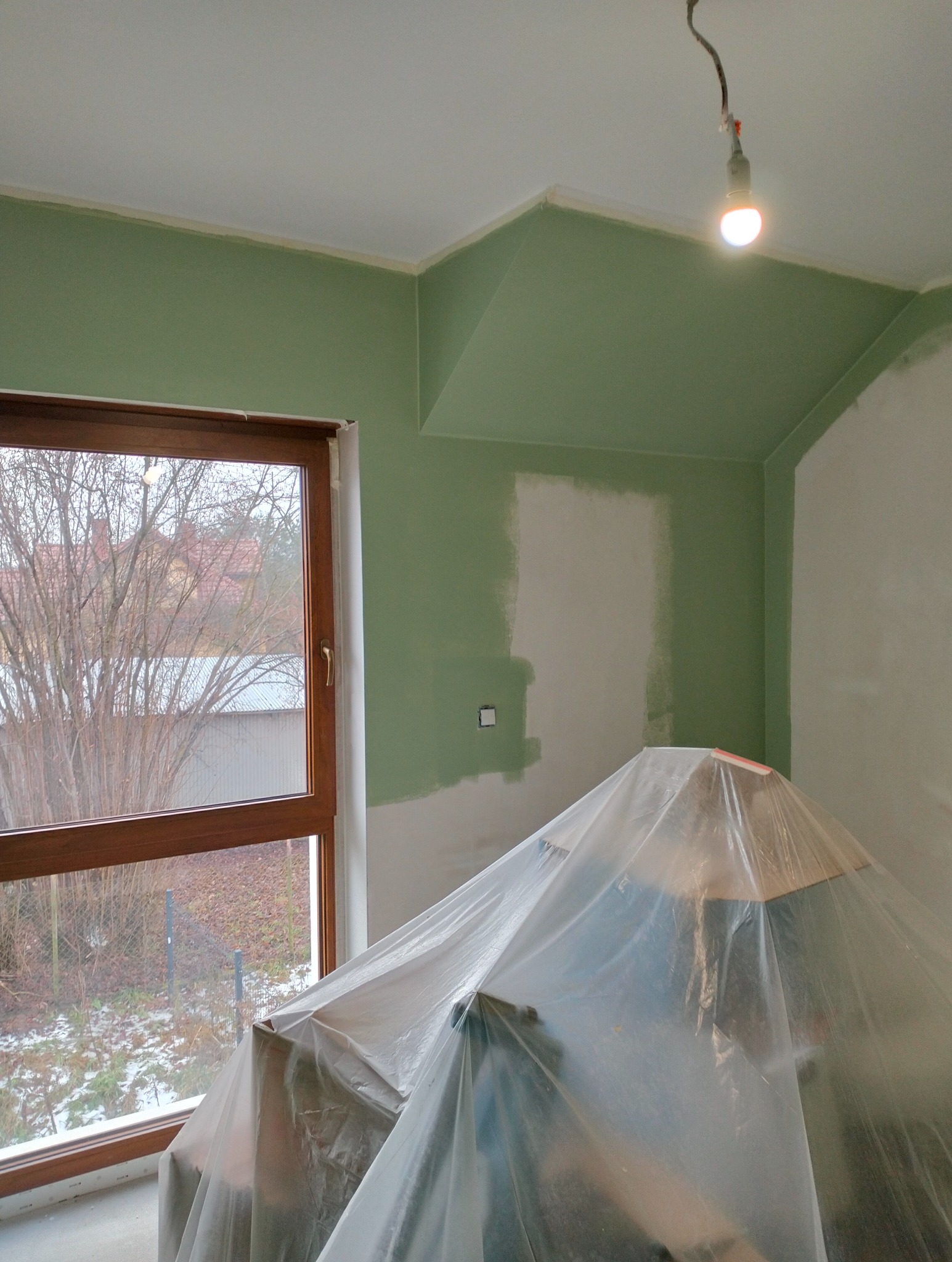 Pokój w trakcie malowania na zielono, z oknem i zabezpieczonym meblem folią. Widoczne ślady szpachlowania i niedomalowane fragmenty ścian. Żarówka oświetla wnętrze.