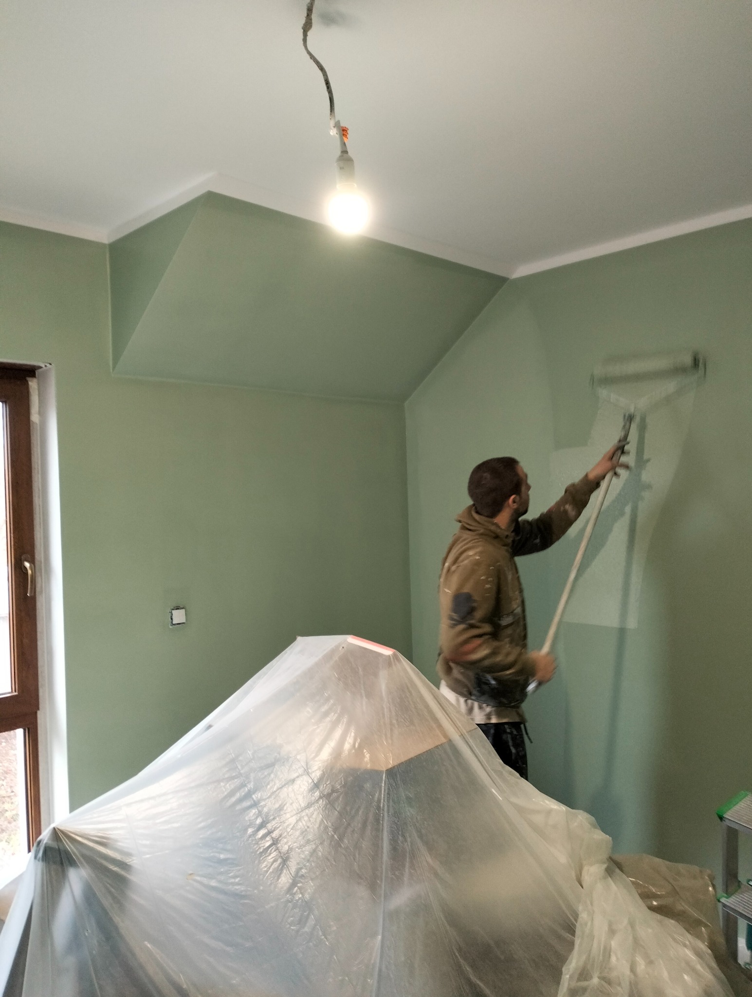 Mężczyzna maluje ścianę na zielono wałkiem. Meble zabezpieczone folią. Widoczna lampa wisząca i okno. Ściany w odcieniu zieleni, sufit biały.