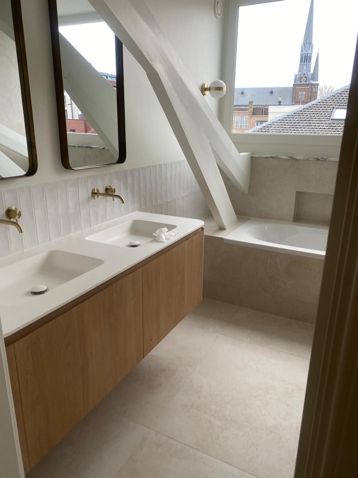 Nowoczesna łazienka na poddaszu w Sompolnie z dwoma umywalkami, drewnianą szafką i widokiem na kościół przez okno. Wykończona w jasnych, neutralnych kolorach.