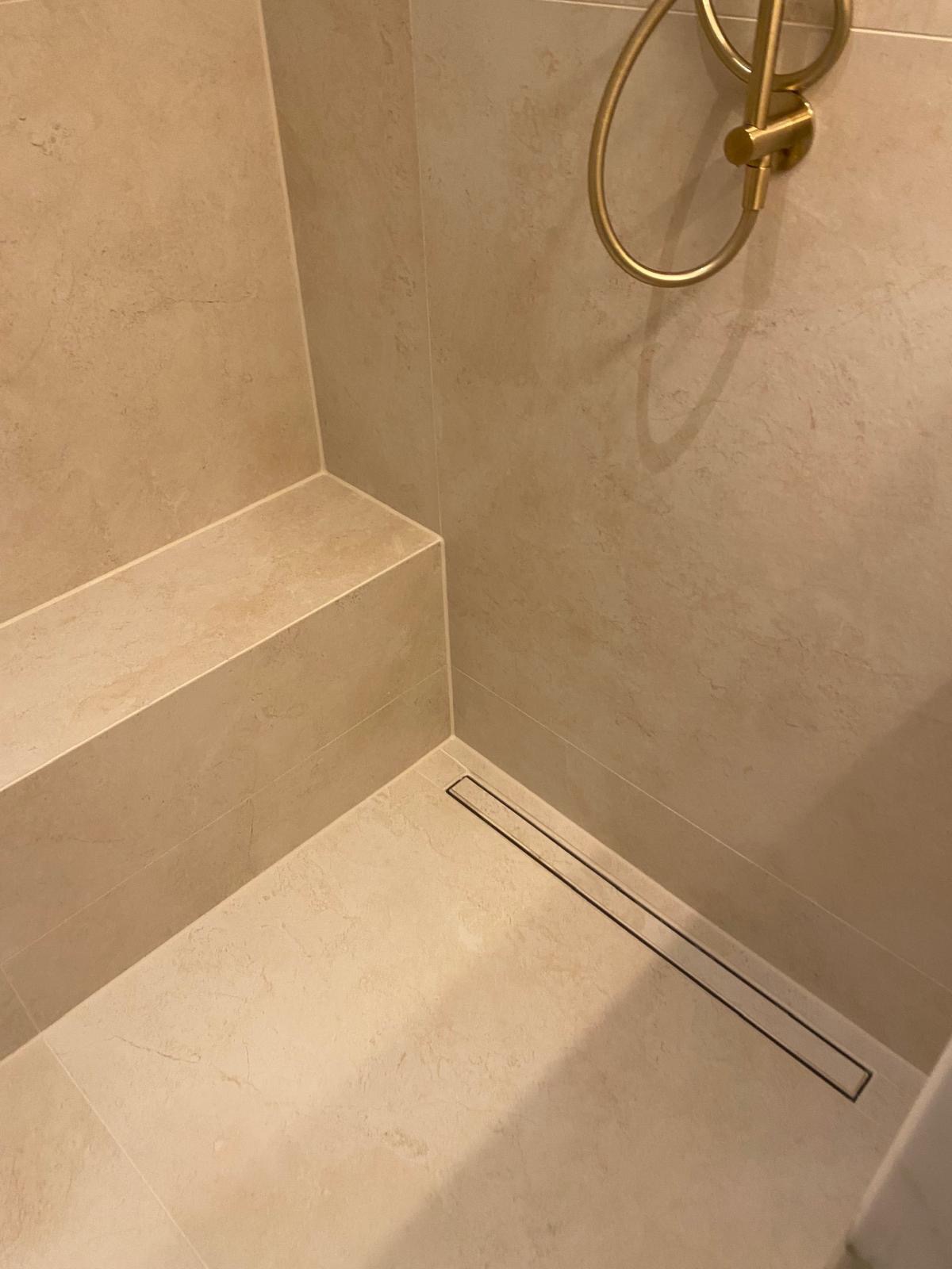 Beżowa kabina prysznicowa z odpływem liniowym i półką z płytek. Złoty uchwyt na ręcznik. Minimalistyczny design, eleganckie wykończenie.