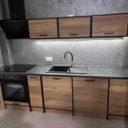 Nowoczesna kuchnia z drewnianymi szafkami, czarnymi ramami i oświetleniem LED podszafkowym. Uwagę zwraca szara ściana z efektem betonu.