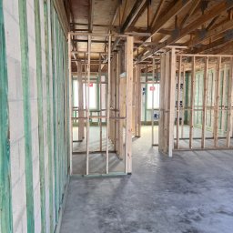 Łukasz Pietkiewicz - Surowy stan budynku szkieletowego: drewniana konstrukcja ścian, stropu i podłogi na betonowej wylewce, widoczne okna. Ściany z zielonymi elementami wzmacniającymi.