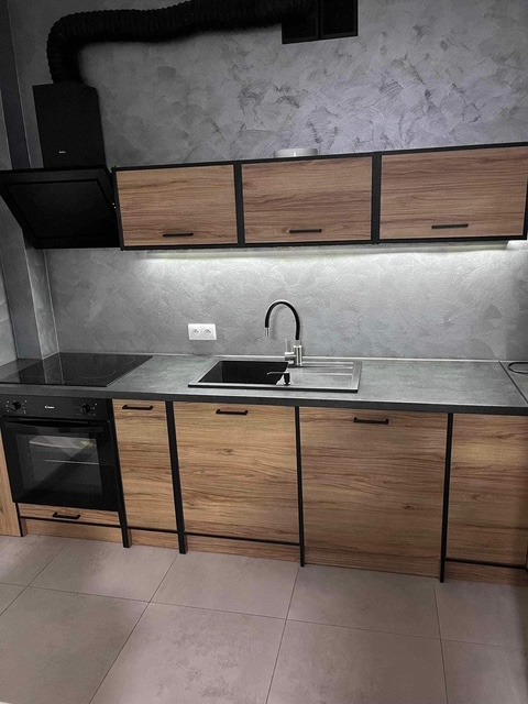 Nowoczesna kuchnia z drewnianymi szafkami, czarnymi ramami i oświetleniem LED podszafkowym. Uwagę zwraca szara ściana z efektem betonu.