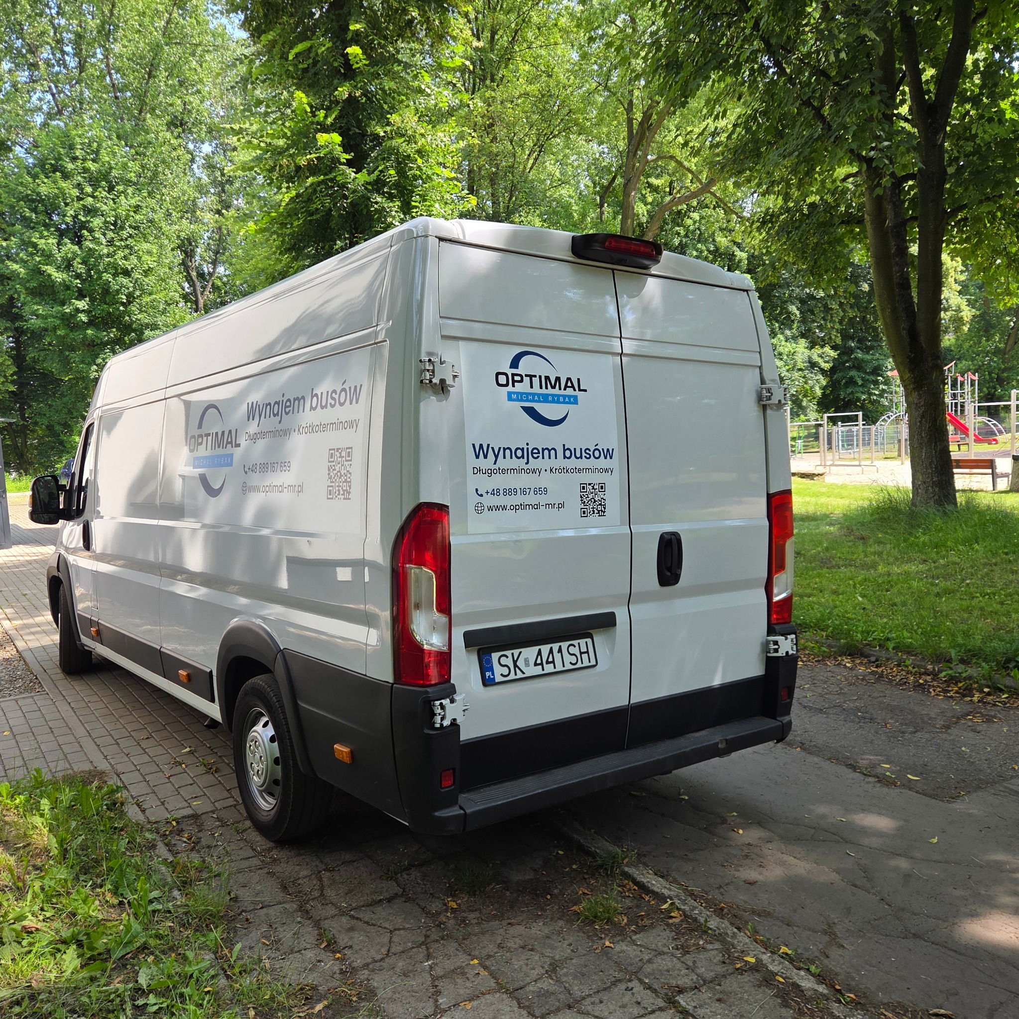 Biały bus firmy Optimal z reklamą wynajmu, zaparkowany na tle zieleni miejskiej. Widoczny numer telefonu i adres strony internetowej.