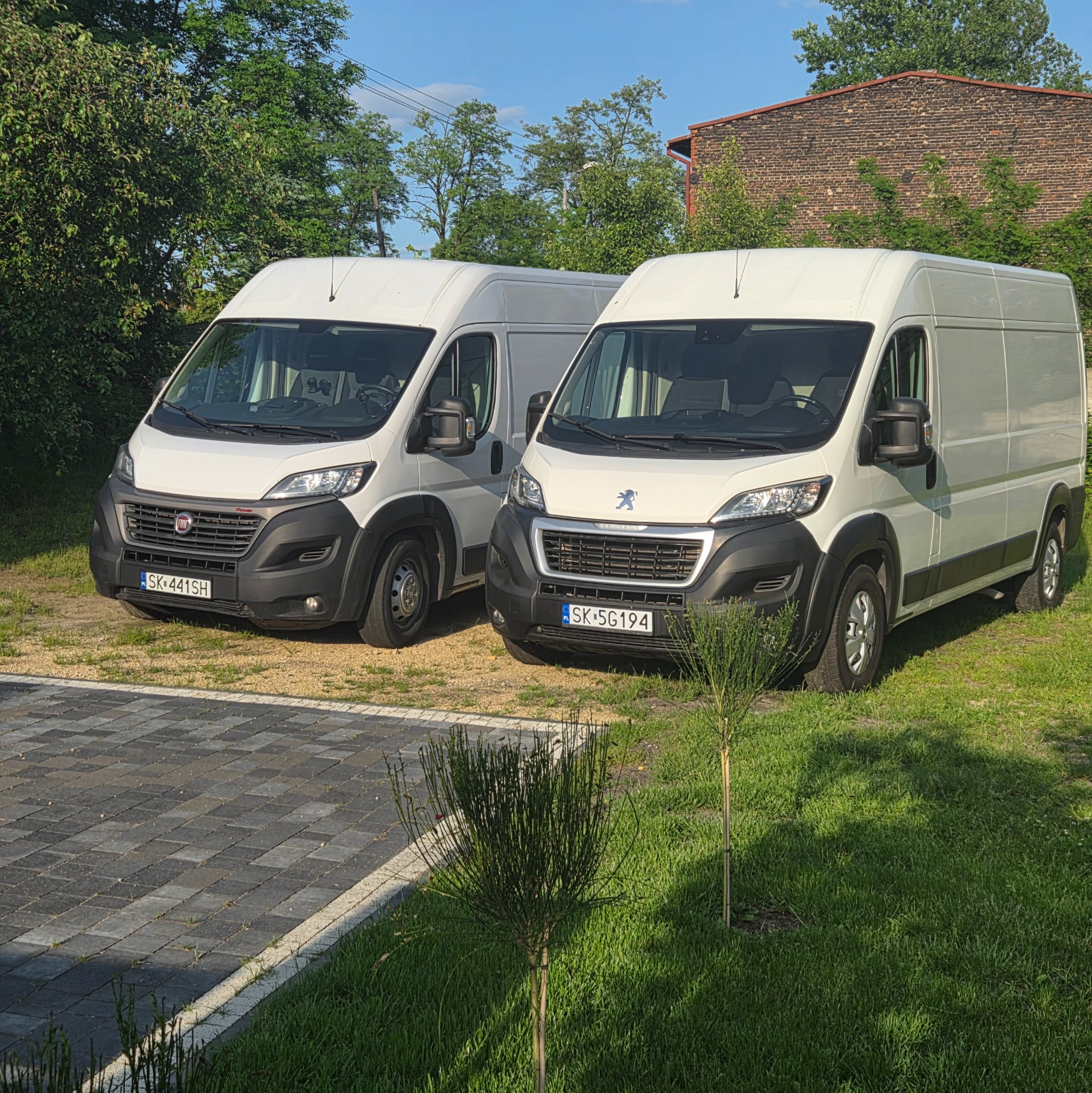 Dwa białe busy: Fiat i Peugeot, stoją obok siebie na trawniku przed budynkiem z czerwonej cegły. Pojazdy z widocznymi tablicami rejestracyjnymi.